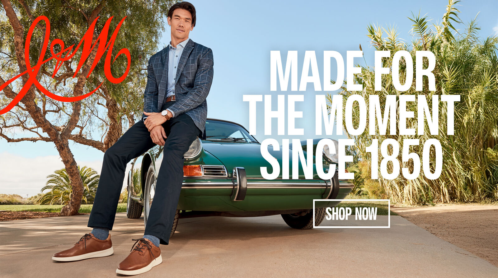 Man leaning on a vintage green car wearing a nicer looking outfit with brown shoes, jeans and a blazer. "Made for the moment since 1850" is written on the right side of the banner.